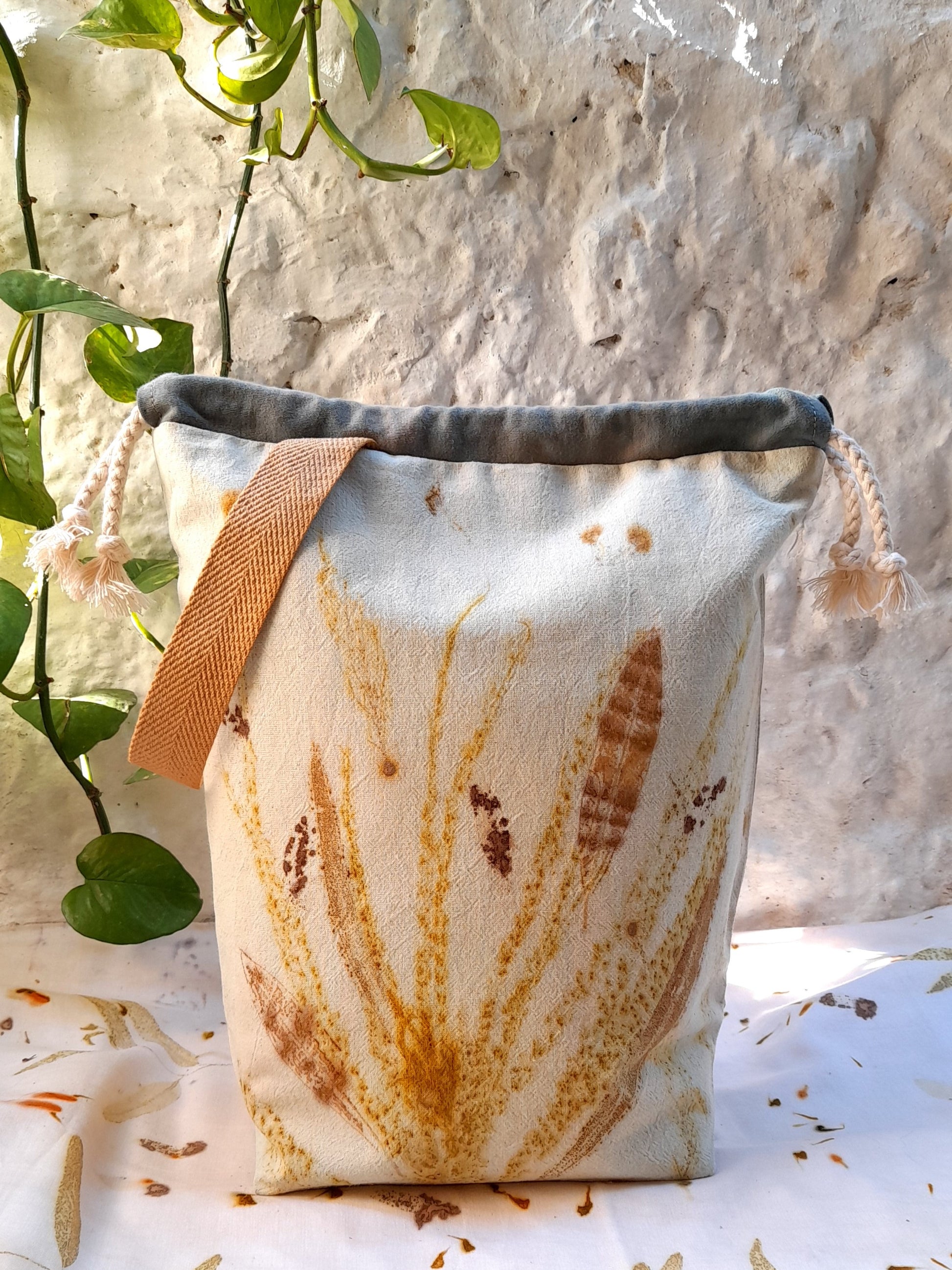 handmade bag for knitters and crocheters, made with ecoprinted fabric in whitecolor and the prints of leaves in various shades of yellow and brown. stanting in front of a white wall with a pothos plant, and over a white table.