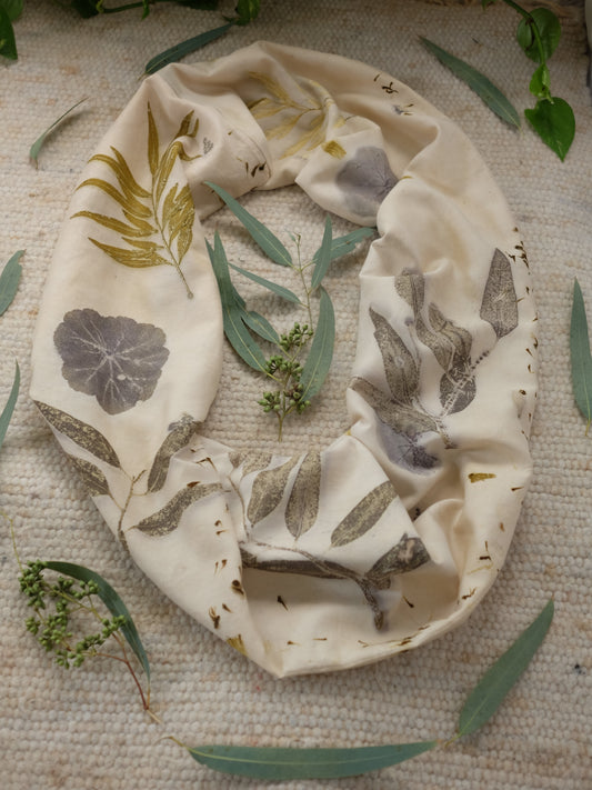 Ecoprinted infinity scarf layed on a carpet, with eucalyptus leaves around it. THe scarf is beige with various leaves printed on it in the colors of ochra, dark grey and brown.