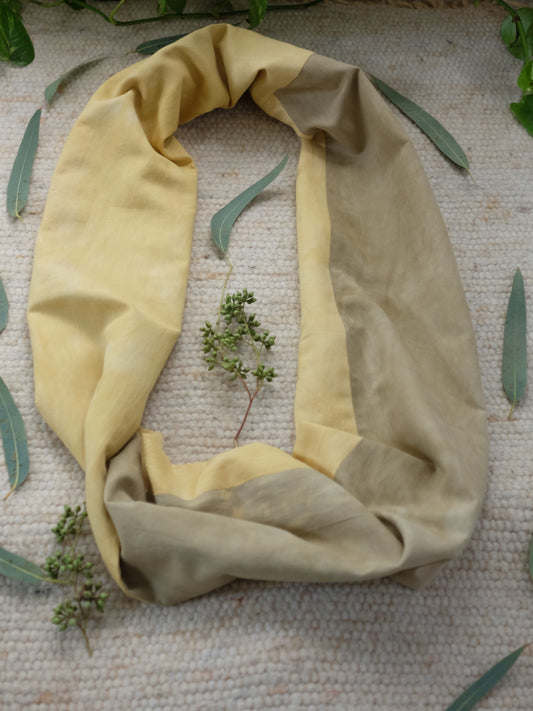 botanically dyed infinity scarf in yellow and khaki color, layed over a white carpet, surrounded withe eucalyptus leaves.