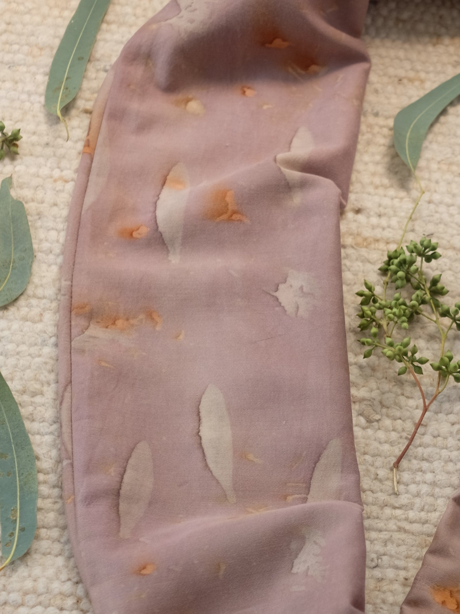section of an ecoprinted scarf , in a grey purple color , surrounded with eucalyptus leaves.