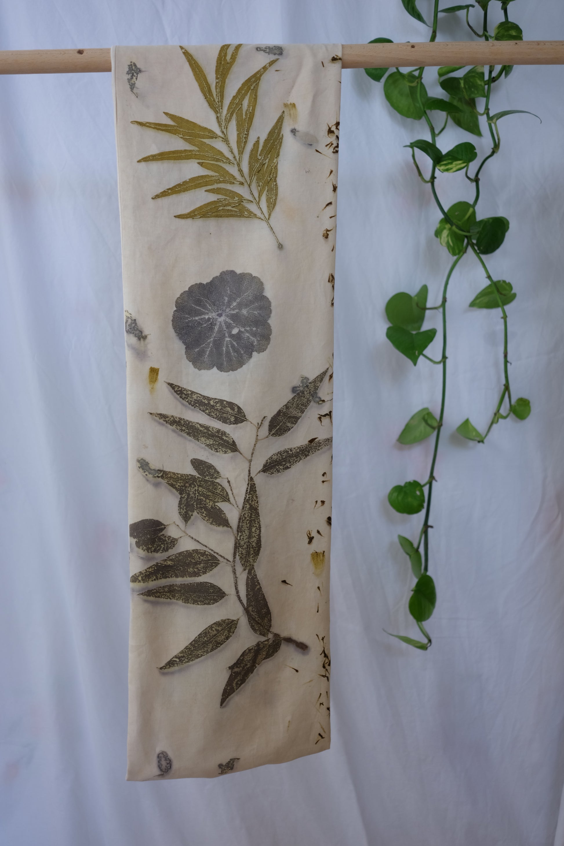cream color ecoprinted infinity scarf, hanging from a dowel. the scarf has ecoprinted eucalyptus, grenvilla and geranious leaves. photographed in a white backround with a pothos plant.