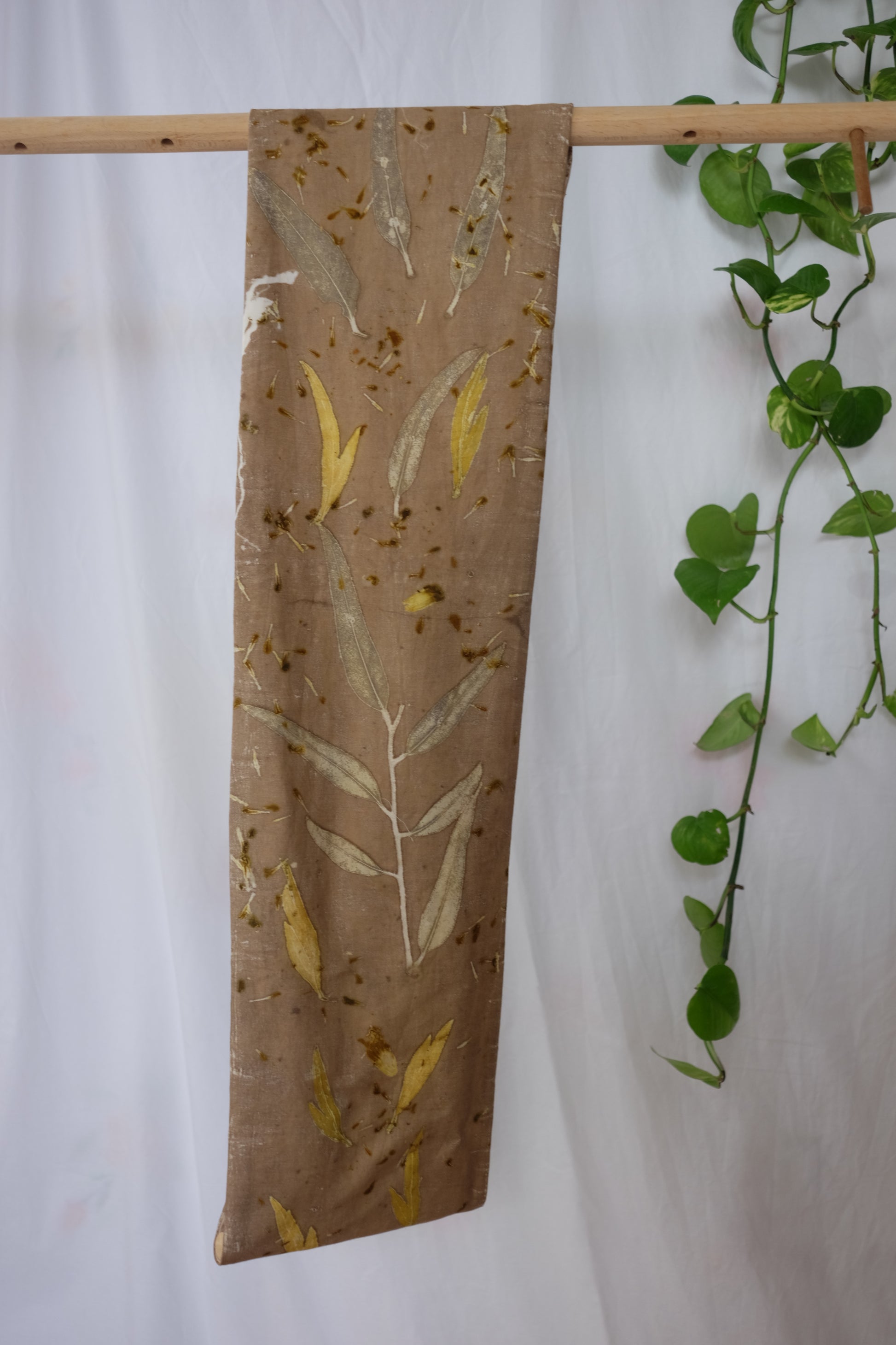 ecoprinted infinity scarf in a khaki color with eucalyptus and grenvila leaves.. Hanging on a wooden dowel with a white backround and a pothos plant in the side.