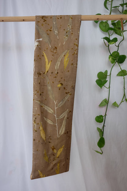 ecoprinted infinity scarf in a khaki color with eucalyptus and grenvila leaves.. Hanging on a wooden dowel with a white backround and a pothos plant in the side.