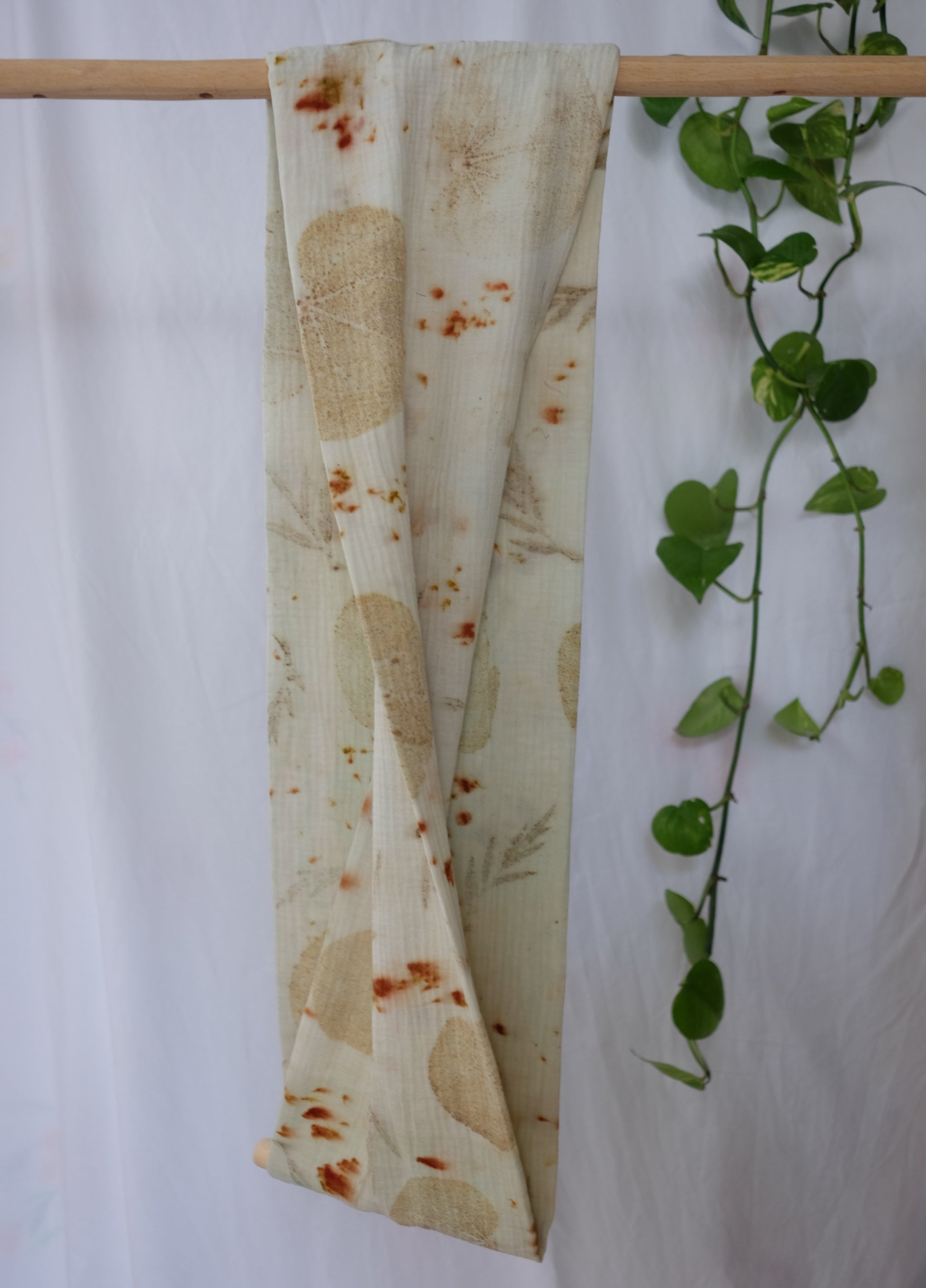 the folded side of the ecoprinted infinity scarf, hanging on a dowel, with a white backround and a pothos pland in the side.