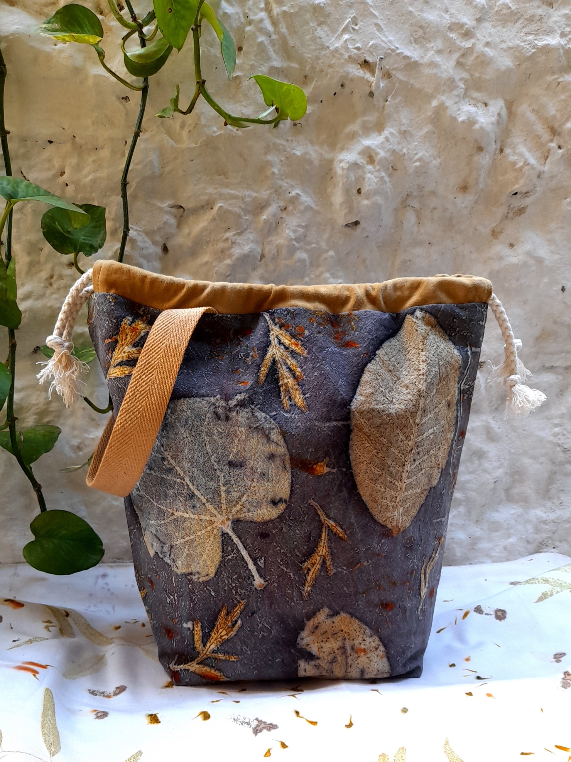 bag handmade with ecoprinted fabric, standing on a table covered with ecoprinted fabric, and in front of a white wall.