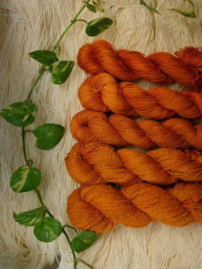 plant based yarns dyed with cosmos flowers in a burn orange color, set over skeins of undeyed yarn with a pothos plant branch on the side.
