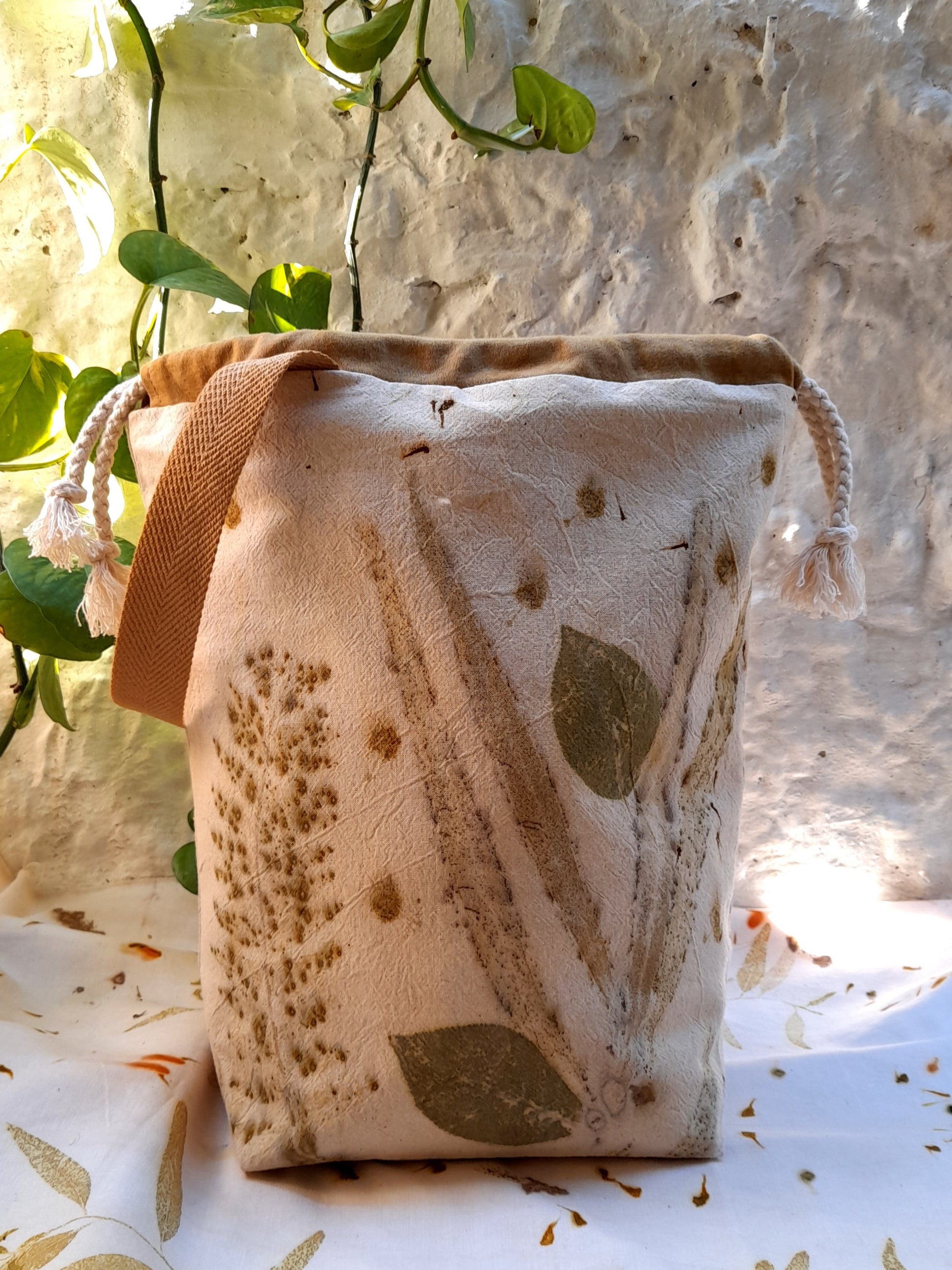 handmade project bag for crocheters and knitters, made with ecoprinted fabric, standing infront of a white wall and on top of a table overed with ecoprinted fabric.