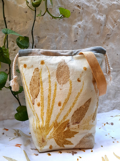 ecoprinted project bag for makers, made with cotton fabric , standing on top of a table covered with ecoprinted fabric and infront of a wall.