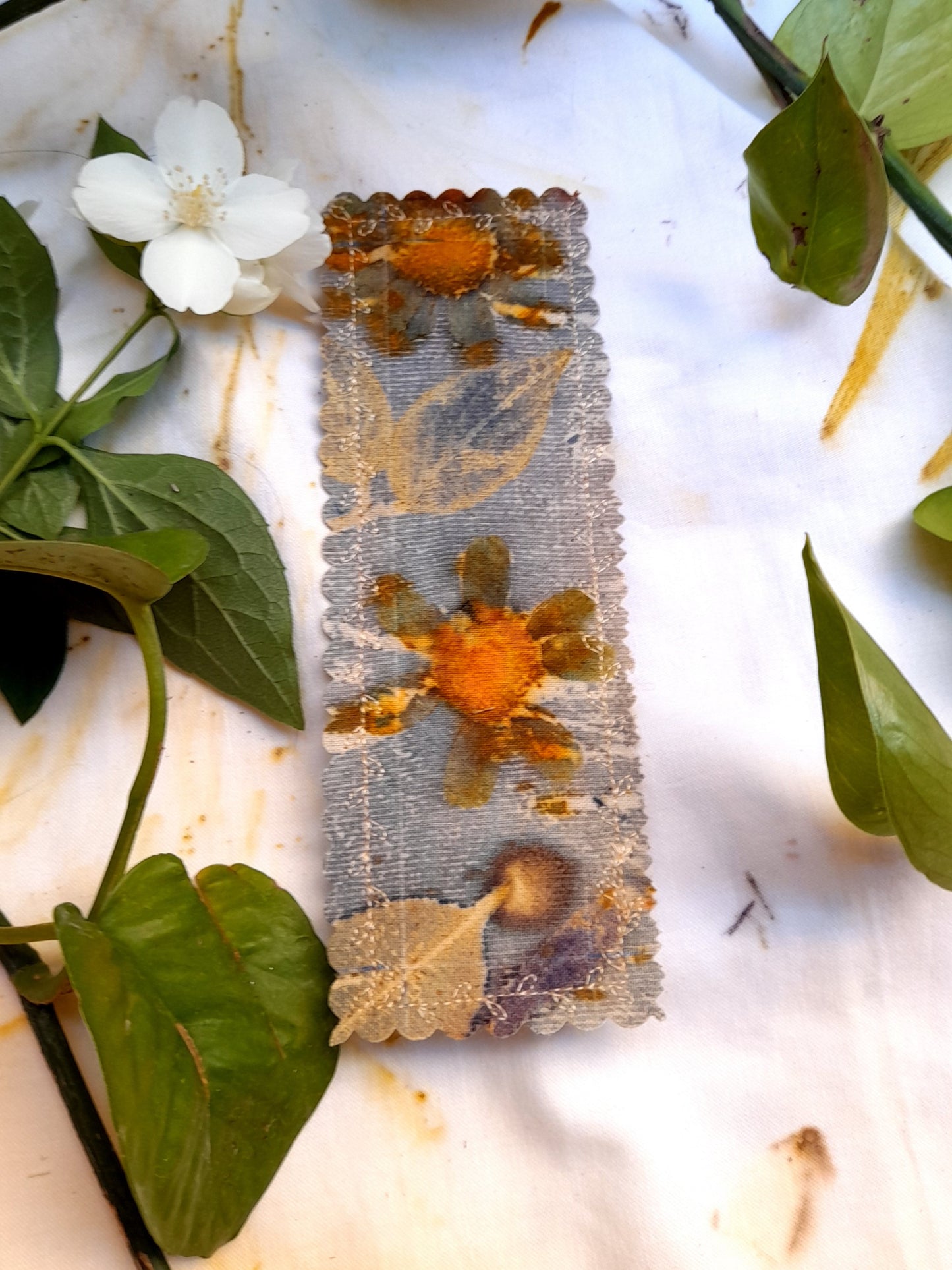 HANDMADE BOOKMARK IN ECOPRINTED COTTON CANVAS, ON A WHITE TABLE WITH PLANTS AROUNT IT