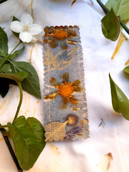 HANDMADE BOOKMARK IN ECOPRINTED COTTON CANVAS, ON A WHITE TABLE WITH PLANTS AROUNT IT